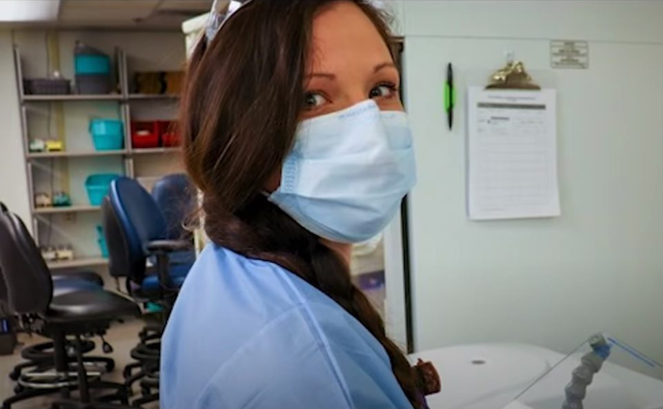 A young woman wears Carolina blue scrubs and a disposable mask. She looks back over her shoulder directly at the camera.