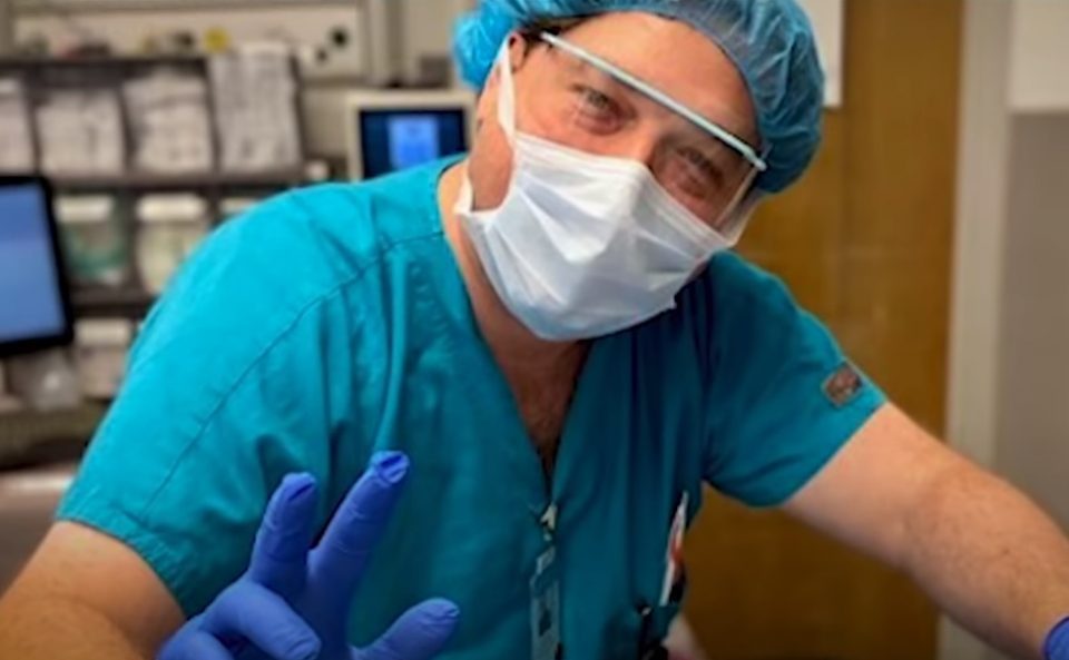 A UNC technician wearing scrubs, protective glasses, and a disposable mask waves at the camera.